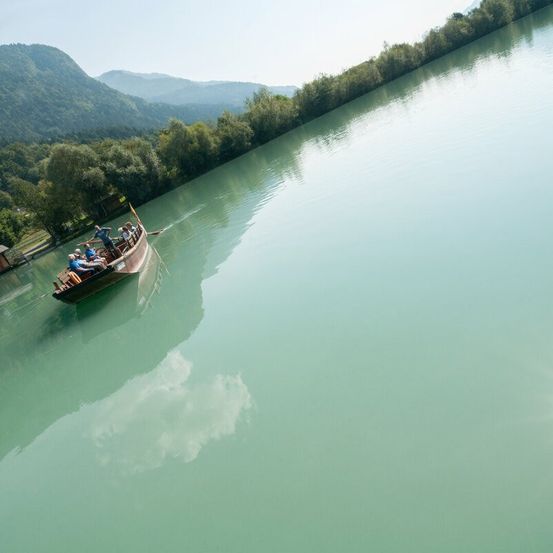 Eine Gruppe von Menschen ist auf einem Boot in einem ruhigen See. Das Boot wird von hinten gepaddelt. Bäume und Berge sind im Hintergrund sichtbar.