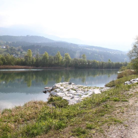 Ein ruhiger See umgeben von üppigem Grün, mit felsigen Ufern und fernen Bergen unter einem klaren Himmel.