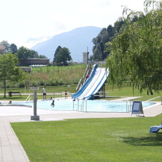 Ein Freibad mit Wasserrutsche ist von üppiger Grünfläche umgeben. Menschen genießen das Becken, einige schwimmen und andere stehen am Rand. Im Hintergrund bilden Berge und Bäume eine ruhige Landschaft.