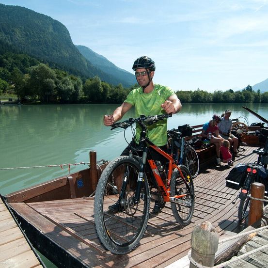 Ein Mann in einem grünen Shirt und einem schwarzen Helm steht auf einem hölzernen Steg und hält ein orangefarbenes Fahrrad. Hinter ihm ist ein kleines Boot festgemacht, und ein Kind sitzt auf dem Steg. Im Hintergrund sind Berge und Bäume zu sehen.