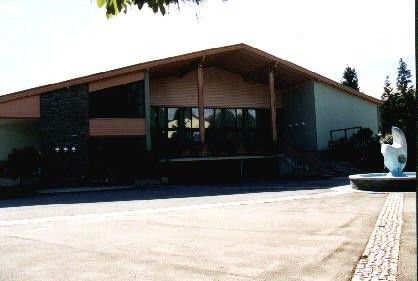 Ein modernes Gebäude mit einem großen Eingang, Glasfenstern und einem Brunnen davor. Es hat ein geneigtes Dach und ist von Bäumen und einer gepflasterten Einfahrt umgeben.