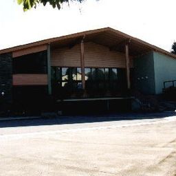 Ein modernes Gebäude mit einem großen Eingang, Glasfenstern und einem Brunnen davor. Es hat ein geneigtes Dach und ist von Bäumen und einer gepflasterten Einfahrt umgeben.