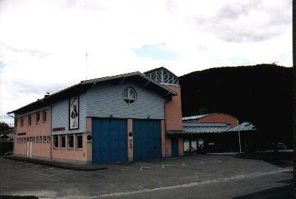 Ein Gebäude mit einer blauen Tür und einem runden Fenster in der Mitte, vor einem großen Berg gelegen. Das Gebäude scheint eine Feuerwache zu sein, mit einem Schild an der Wand.