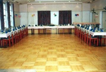 Ein leerer Bankettsaal mit langen Tischen und Stühlen, weißen Tischdecken und bunten Luftballons. Der Boden ist aus Holz, und der Raum hat große Fenster und Lampen an der Decke.