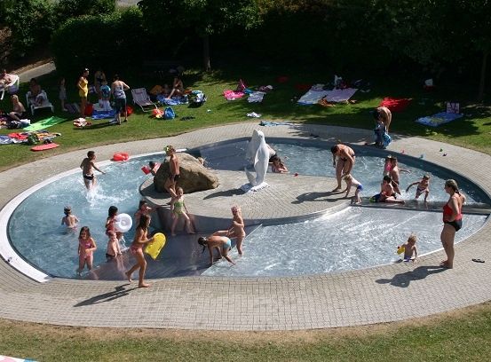 Eine Luftaufnahme eines großen Außenpools mit schwimmenden Kindern und einer weißen Statue. Leute entspannen sich auf dem Gras um den Pool herum.