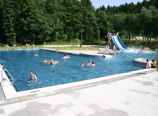 Ein großer Außenpool mit Schwimmern und einer Wasserrutsche. Menschen sitzen auf der Wiese und einige auf der Terrasse.