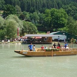 Eine Gruppe von Menschen befindet sich auf einem Boot auf einem Fluss, mit einer Person, die vorne steht. Das Boot hat Blumen darauf. In der Ferne sind mehrere Menschen, Bäume und ein Gebäude zu sehen.