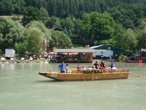 Eine Gruppe von Menschen befindet sich auf einem Boot auf einem Fluss, mit einer Person, die vorne steht. Das Boot hat Blumen darauf. In der Ferne sind mehrere Menschen, Bäume und ein Gebäude zu sehen.