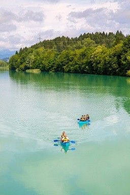 Drei Kajakfahrer paddeln auf einem türkisfarbenen See, umgeben von üppigem Grün, unter einem teilweise bewölkten Himmel.