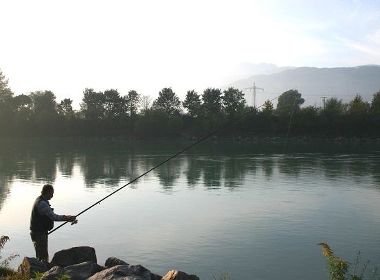 Ein Mann steht mit einer Angelrute am Fluss, umgeben von Bäumen und Bergen.