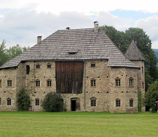 Ein Steingebäude mit einer Holztür steht auf einer großen Wiese, mit Bäumen und Bergen im Hintergrund.