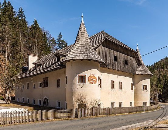 Ein großes, beigefarbenes Gebäude mit einem spitzen Dach steht in einer ländlichen Umgebung, umgeben von einem Holzzaun. Im Hintergrund sind Bäume und Berge zu sehen.