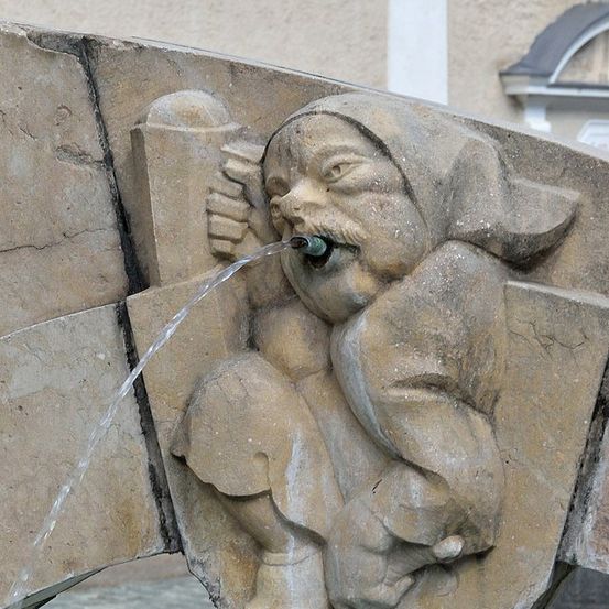 Eine Steinbrunnenskulptur zeigt einen Gnom, aus dessen Mund Wasser fließt. Sie ist an einem Steinbogen befestigt, mit einem Gebäude im Hintergrund.
