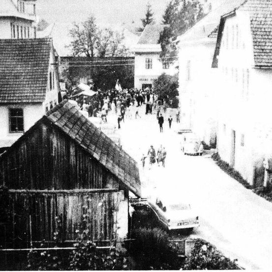 Ein Schwarz-Weiß-Foto zeigt eine Straße mit mehreren Menschen, die sich um ein Gebäude mit Bäumen im Hintergrund versammelt haben.