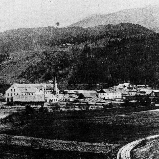 Ein Schwarz-Weiß-Foto zeigt eine kleine Stadt, die in einem Tal mit Bergen im Hintergrund eingebettet ist. Es gibt mehrere Gebäude, darunter eine prominente Fabrik. Das Gebiet ist von Bäumen und Feldern umgeben.