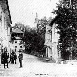 Ein Schwarz-Weiß-Foto von 1920 zeigt eine schmale Straße mit mehreren stehenden und plaudernden Personen. Die Gebäude auf beiden Seiten haben Vordächer, und es sind Bäume und eine Kirche in der Ferne zu sehen.