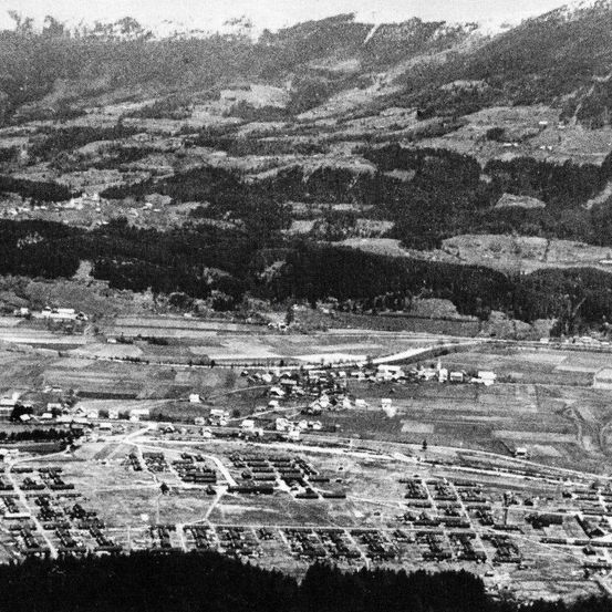 Eine Luftaufnahme einer Stadt, die von Bergen und Wäldern umgeben ist. Die Stadt hat viele Gebäude, von denen einige im Bau sind. Die Stadt ist von einem großen Landgebiet umgeben.