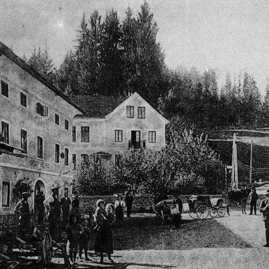 Ein Schwarz-Weiß-Foto zeigt Menschen vor einem Gebäude stehend. Im Vordergrund steht eine Kutsche mit Pferd, die von einer Person gepflegt wird. Bäume und weitere Gebäude sind im Hintergrund zu sehen.