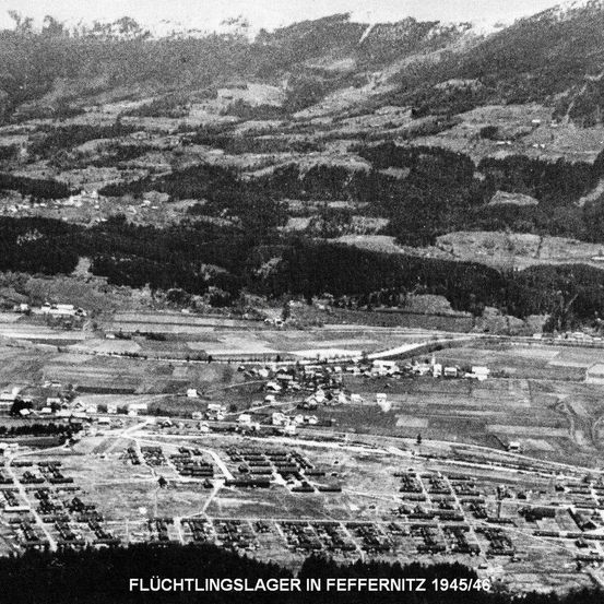 Eine Schwarz-Weiß-Luftaufnahme des Flüchtlingslagers in Feffernitz, Österreich, von 1945/46. Das Lager ist von dichten Wäldern und hügeligem Gelände umgeben. Viele Gebäude sind sichtbar, einige in einem Zustand der Verwahrlosung.