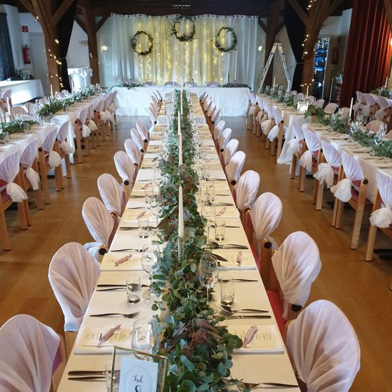Ein Bankettsaal mit langen Tischen, die mit weißen Tüchern bedeckt und mit Grünpflanzen und Kerzen dekoriert sind. Die Stühle sind mit weißem Stoff bezogen und in Reihen angeordnet. Im Hintergrund befindet sich ein weißer Vorhang mit goldenen Lichtern und einem Feuerlöscher.
