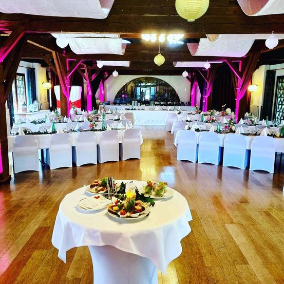 Ein Bankettsaal mit Holzboden und pinker Beleuchtung. Viele Tische sind mit weißen Stühlen und Tischdecken gedeckt. Jeder Tisch hat eine Tischdekoration und dekorative Teller.