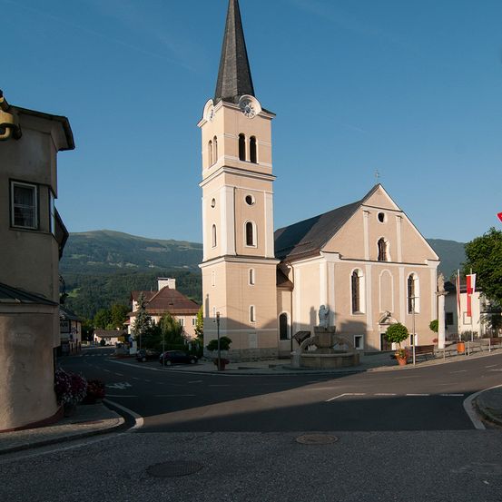 Eine Kirche mit hohem Turm und Uhr steht in einer Stadt. Vor ihr befindet sich ein Brunnen, mit Bergen im Hintergrund.