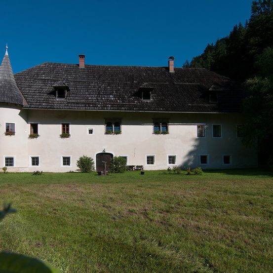 Ein großes weißes Gebäude mit grauem Dach und spitzen Türmen, umgeben von Grün und Bäumen.