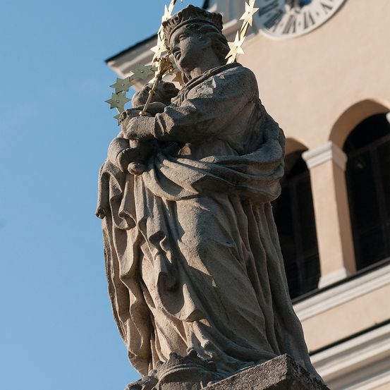 Eine Statue einer gekrönten Frau, die ein Baby und einen sternbesetzten Stab hält, steht vor einem Gebäude mit Bogenfenstern und einem Uhrturm.
