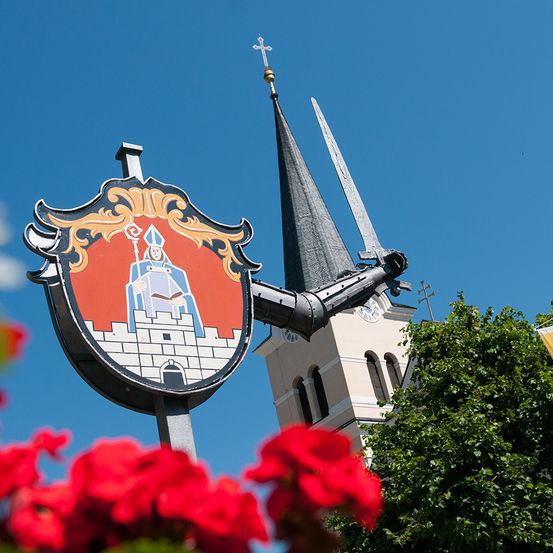Ein Wappenschild mit einer gekleideten Figur, die einen Stab und ein Buch hält, steht vor einem Kirchturm. Die Kirche ist weiß mit einem Kreuz oben und einer Uhr an der Seite.