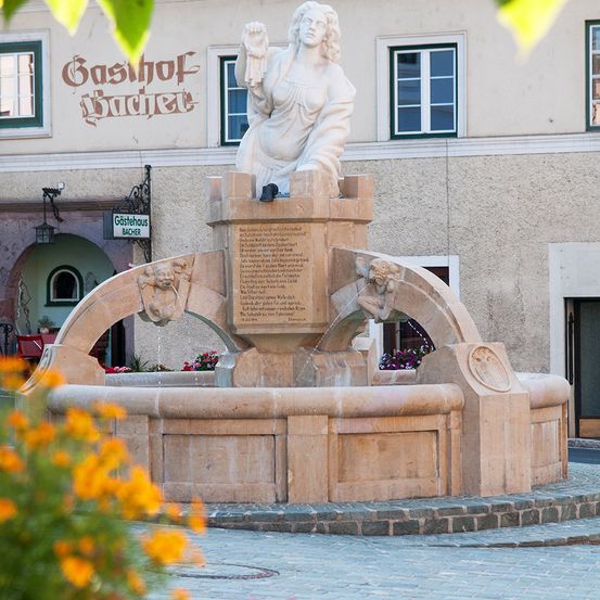Ein Steinbrunnen mit einer Statue einer Frau oben steht in der Mitte einer Kopfsteinpflasterstraße. Der Brunnen hat eine Inschrift auf seiner Basis. Dahinter befindet sich ein Gebäude mit Fenstern und einem Schild, das Gasthof Bacher besagt.