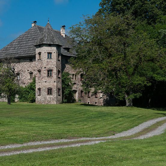 Ein altes Steingebäude mit einem spitzen Dach und Schornsteinen steht auf einem Rasenfeld. Bäume umgeben das Gebäude, einer davon im Vordergrund. Der Himmel ist blau mit einigen Wolken.