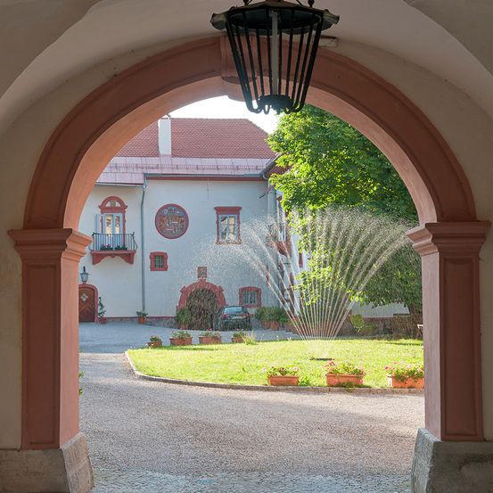 Ein viktorianischer Bogen führt zu einer Einfahrt mit einem Springbrunnen im Garten. Das Haus dahinter hat ein rotes Dach und rot umrandete Fenster. Bäume und Pflanzen umgeben den Garten.