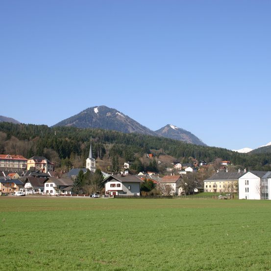Ein Dorf mit mehreren Häusern, einer Kirche und einem breiten grünen Feld liegt unterhalb bewaldeter Berge unter einem klaren blauen Himmel.