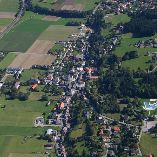 Luftaufnahme eines Dorfes mit verstreuten Häusern, grünen Feldern und Bäumen. Das Dorf ist von üppigen grünen Landschaften umgeben. In der Ferne ist ein blauer Swimmingpool zu sehen.