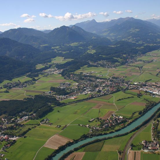 Luftaufnahme eines grünen Tals mit Feldern, Bergen und einem mäandrierenden Fluss unter teilweise bewölktem Himmel.