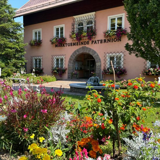 Ein rosafarbenes Gebäude mit der Aufschrift 'Marktgemeinde Paternion' hat Blumen an seinen Fenstern. Der Eingang hat einen Bogen. Vor dem Gebäude befindet sich ein Garten mit einer Vielzahl von Blumen.