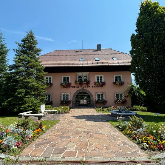 Ein großes rosa Gebäude mit vielen Fenstern und Blumen davor, beschriftet mit 'MARKTGEBMEINDE PATERION'. Ein Steinweg führt zum Eingang und ist von Bänken flankiert und von üppiger Grünfläche umgeben.