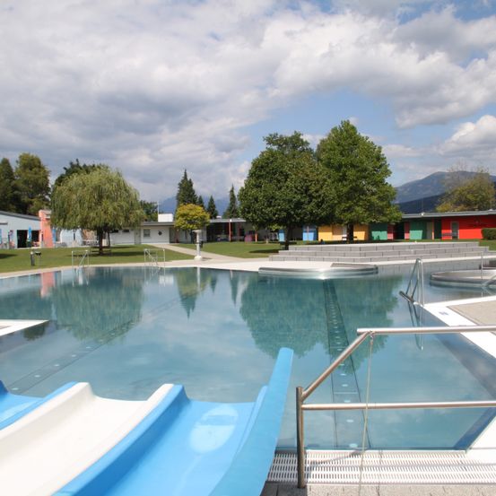 Ein Freibad mit einer Rutsche und Sprungbrett unter einem teilweise bewölkten Himmel, umgeben von Bäumen und Gebäuden.