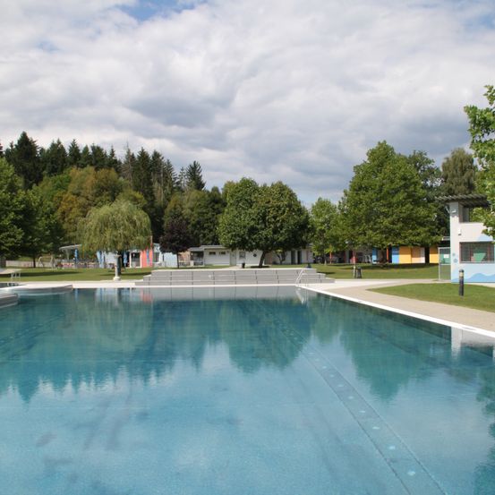 Ein leerer Außenpool mit klarem blauem Wasser, umgeben von einer Rasenfläche mit Bäumen und Gebäuden.