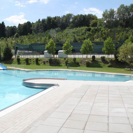 Ein leerer Außenpool mit einer Rutsche links und einem Terrassenbereich davor. Grünflächen und Bäume umgeben das Areal.