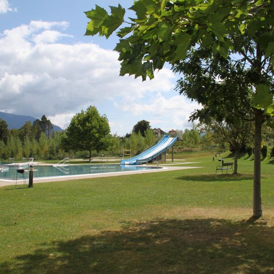 Ein Freibad mit einer blauen Rutsche, umgeben von einer Rasenfläche und Bäumen, mit einem klaren Himmel und Wolken im Hintergrund.