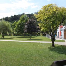 Ein Park mit einigen Bäumen und Bänken. Die Bäume sind in voller Blüte, und der Himmel ist blau mit einigen Wolken. In der Ferne befindet sich ein Gebäude mit einem Zaun.