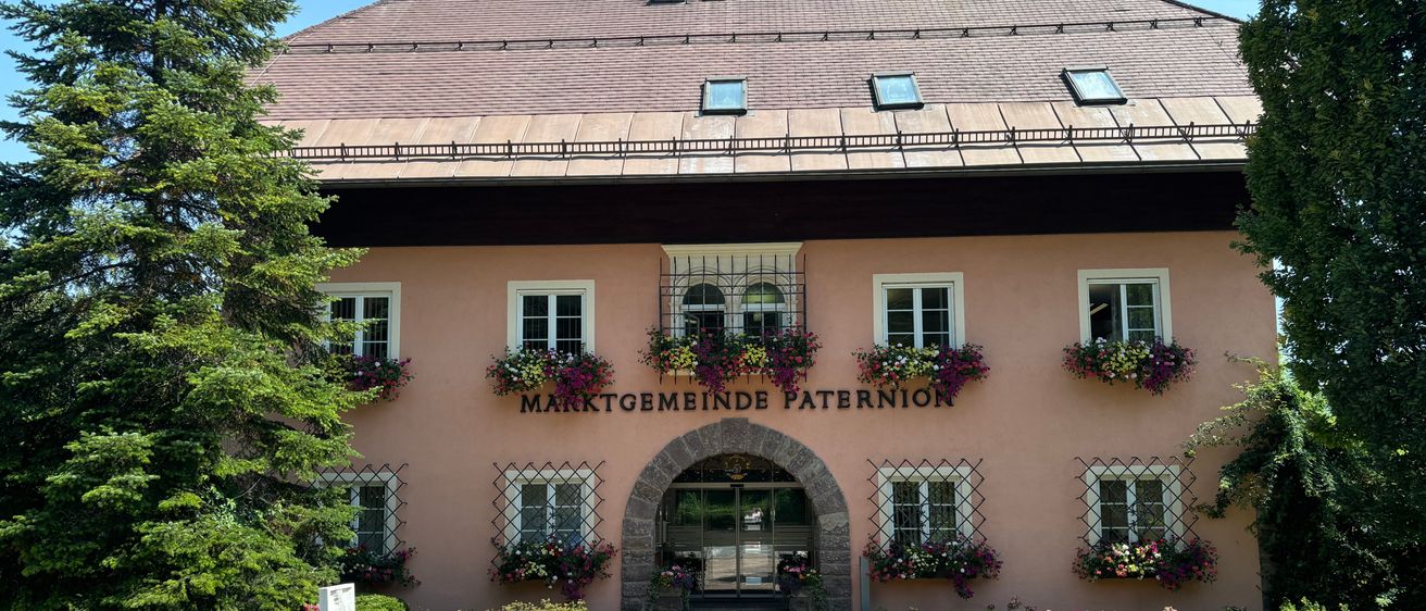 Ein rosafarbenes Gebäude mit einem braunen Dach und einem gewölbten Eingang. Das Gebäude hat mehrere Fenster, die mit Blumenkästen geschmückt sind. Darüber steht 'MARKTGEMEINDE PATERION'. Das Gebäude ist von einem Garten mit verschiedenen Blumen umgeben.