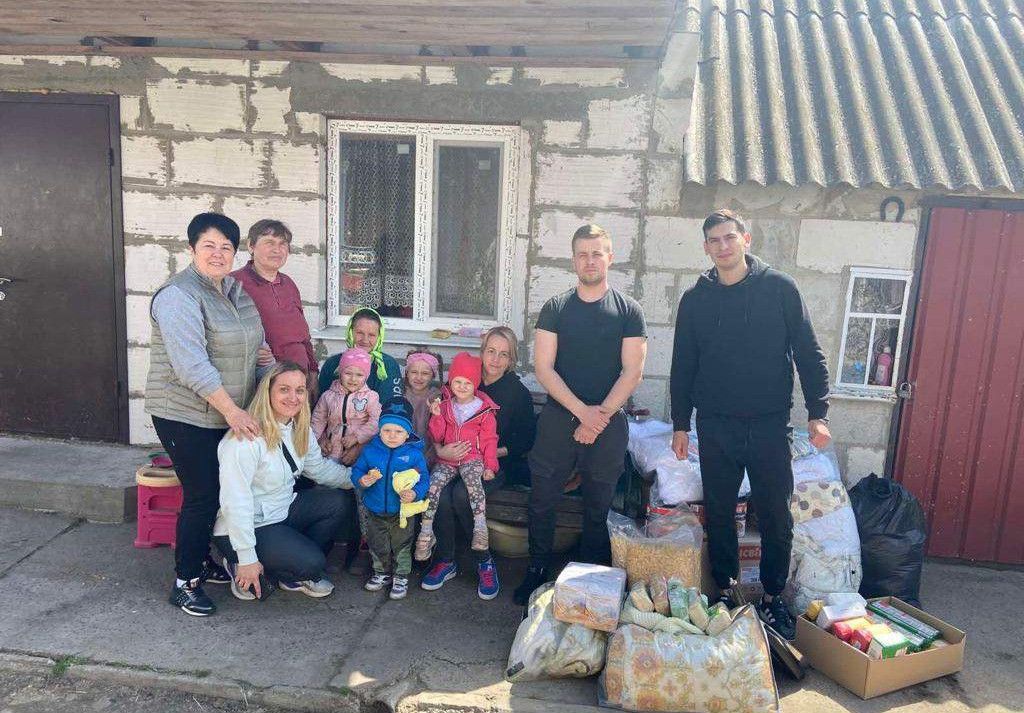 Eine Gruppe von Menschen steht vor einem Haus mit Säcken voller Vorräte. Zwei Männer in Schwarz stehen rechts. Zwei Frauen hocken vor Kindern.