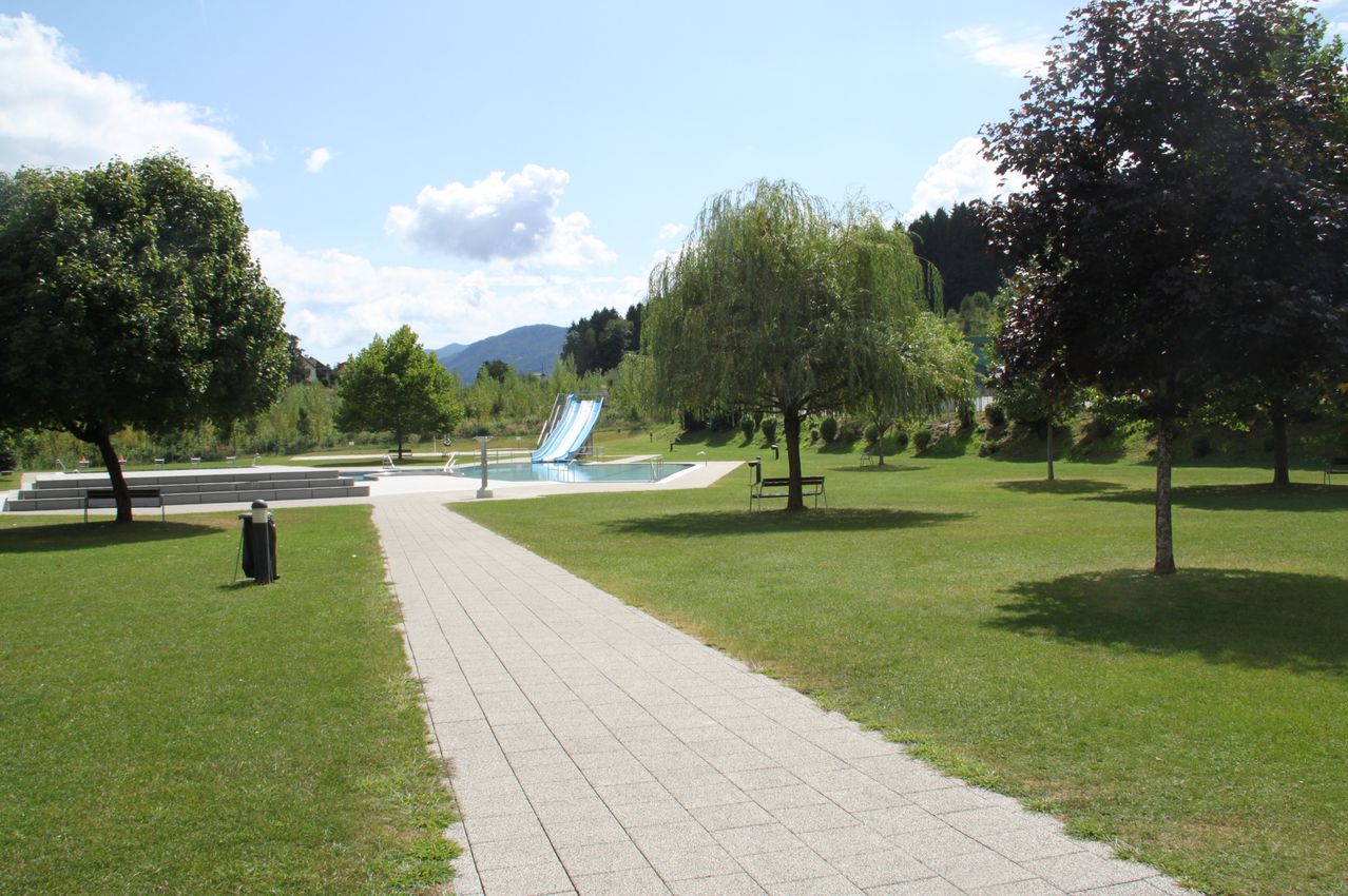 Ein ruhiger Park mit einem gepflasterten Weg, der zu einem Schwimmbecken mit Wasserrutsche führt, umgeben von saftigem grünem Rasen und Bäumen, unter einem teilweise bewölkten Himmel.