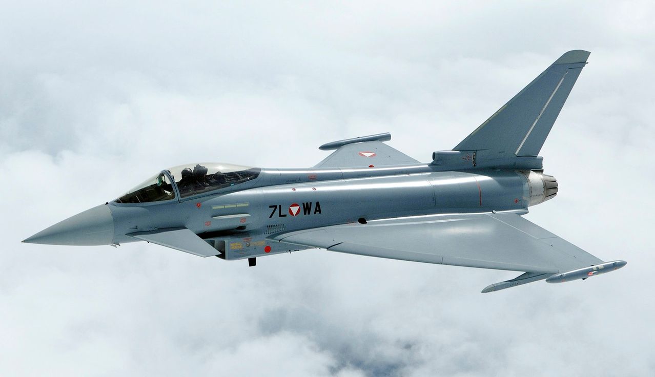 Ein graues Kampfflugzeug mit 7L OWA an der Seite fliegt durch die Wolken. Die Cockpit ist sichtbar, mit Piloten im Inneren.