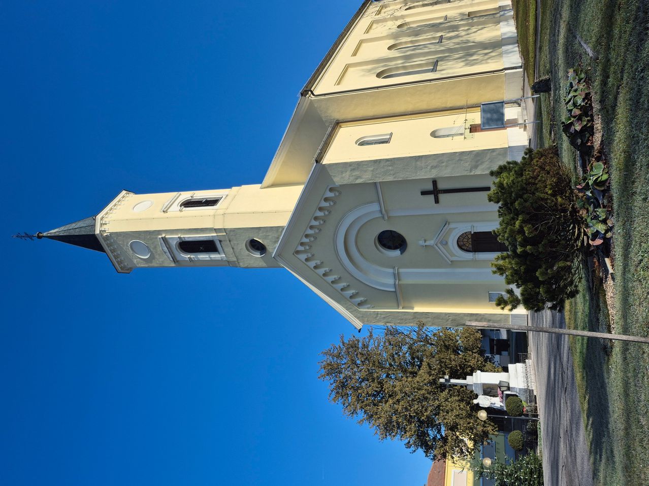 Eine weiße Kirche mit einem Turm, einem Kreuz über dem Eingang und mehreren Rundbogenfenstern unter einem klaren blauen Himmel.