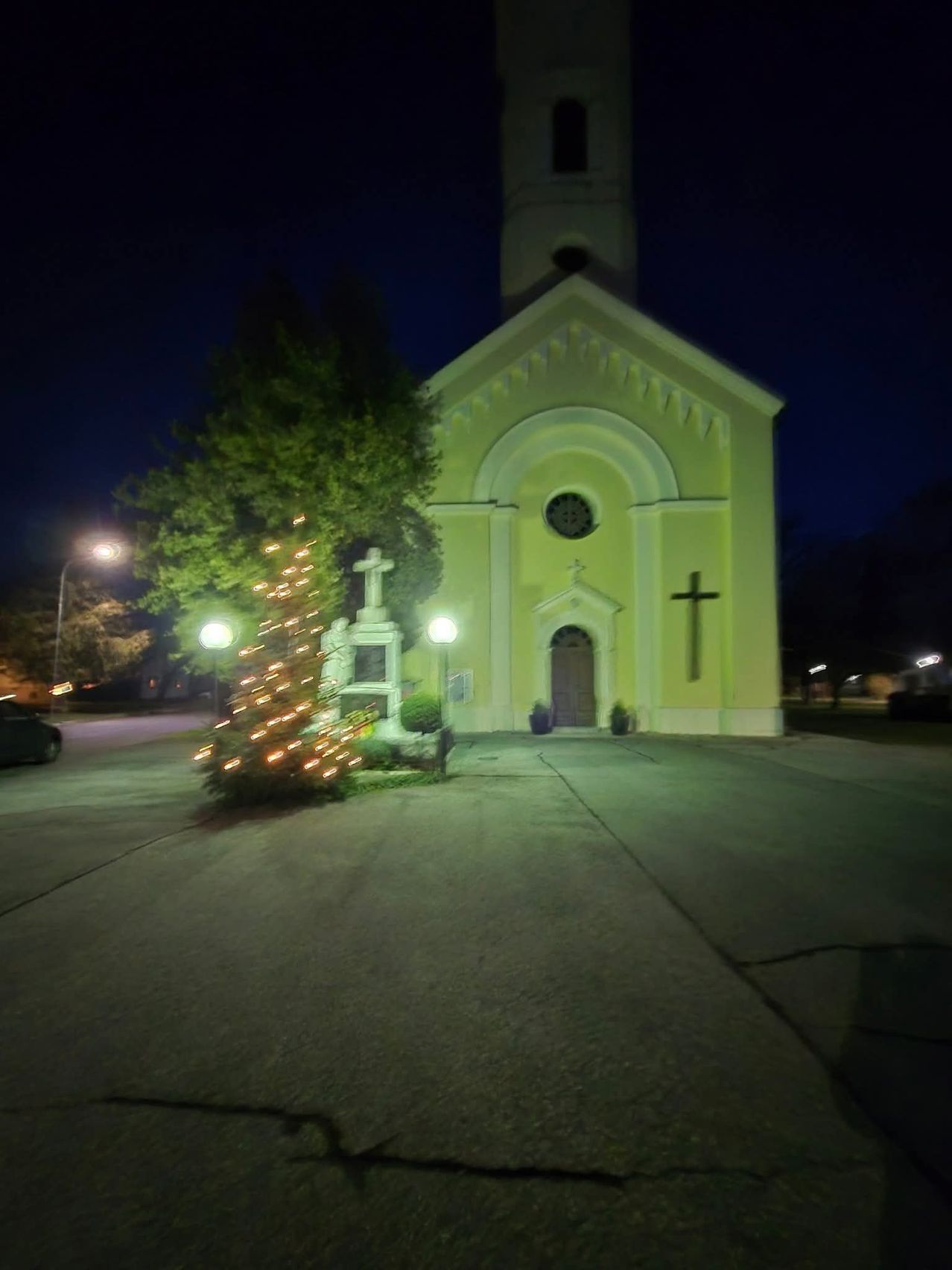 Eine hell beleuchtete Kirche bei Nacht mit einem geschmückten Weihnachtsbaum davor, einem Kreuz an der Wand und zwei Topfpflanzen vor der Tür.