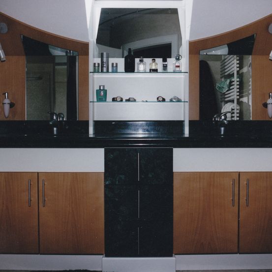 Ein Badezimmer mit einem großen Spiegel, Regalen, zwei Waschbecken und Holzschränken. Die Regale haben verschiedene Flaschen und Gegenstände.