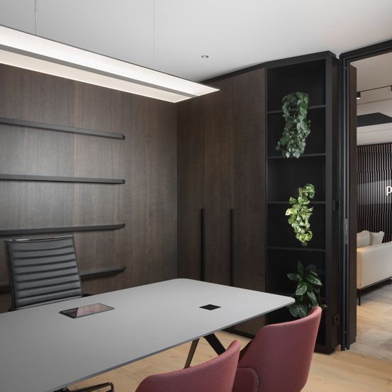 Ein Büroraum mit Holzwänden, einem weißen Tisch und roten Stühlen. Ein schwarzer Regal mit Pflanzen steht rechts, und ein weißes Sofa ist durch eine offene Tür sichtbar.